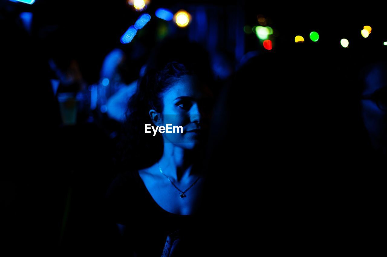 Young woman looking away while standing against illuminated lights at night