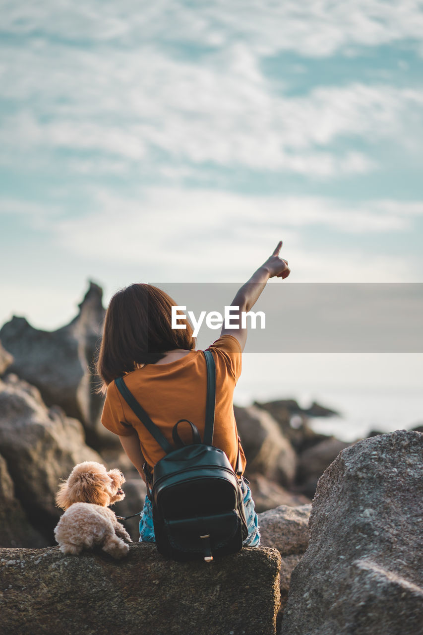 Rear view of woman sitting with dog on rocky shore against sky