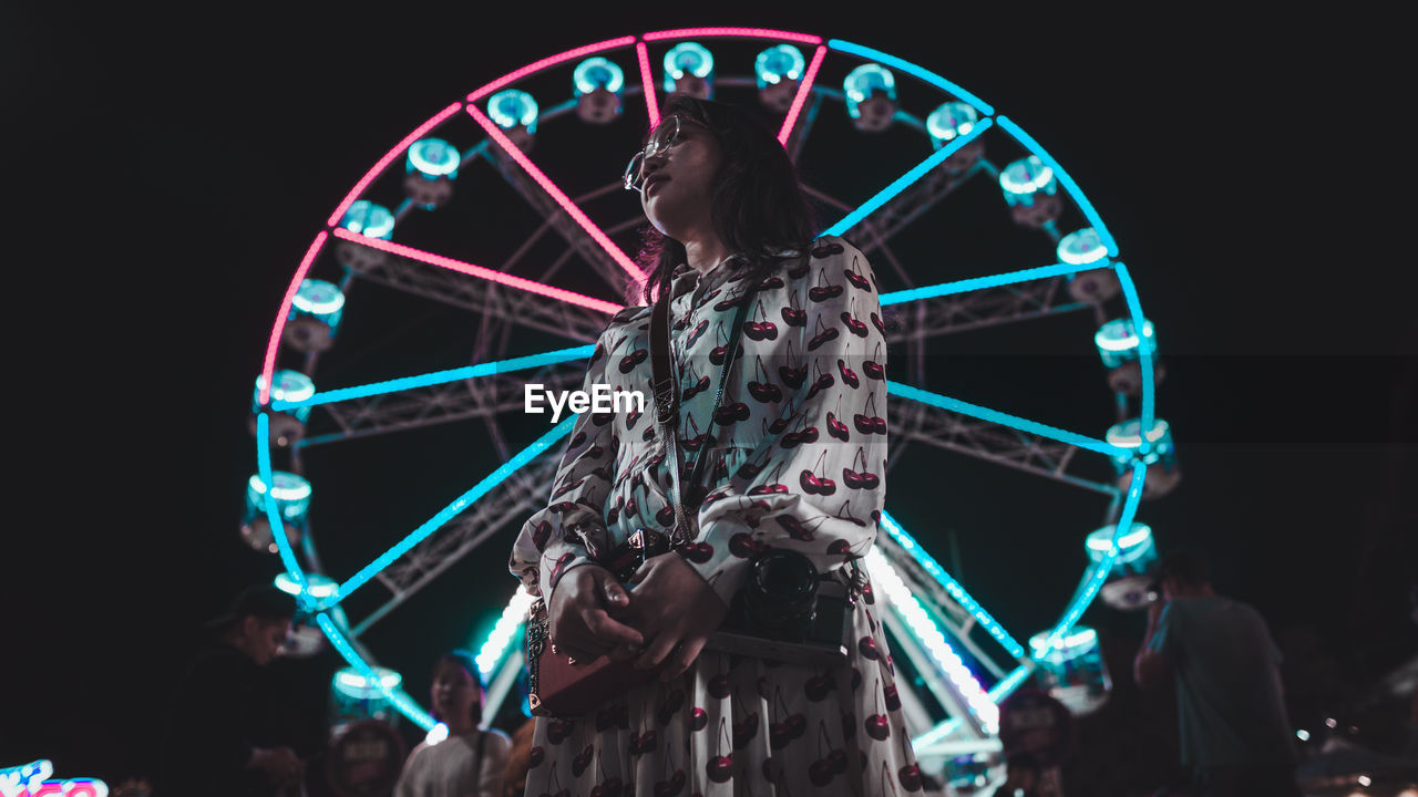 Low angle view of woman standing against ferris wheel at night