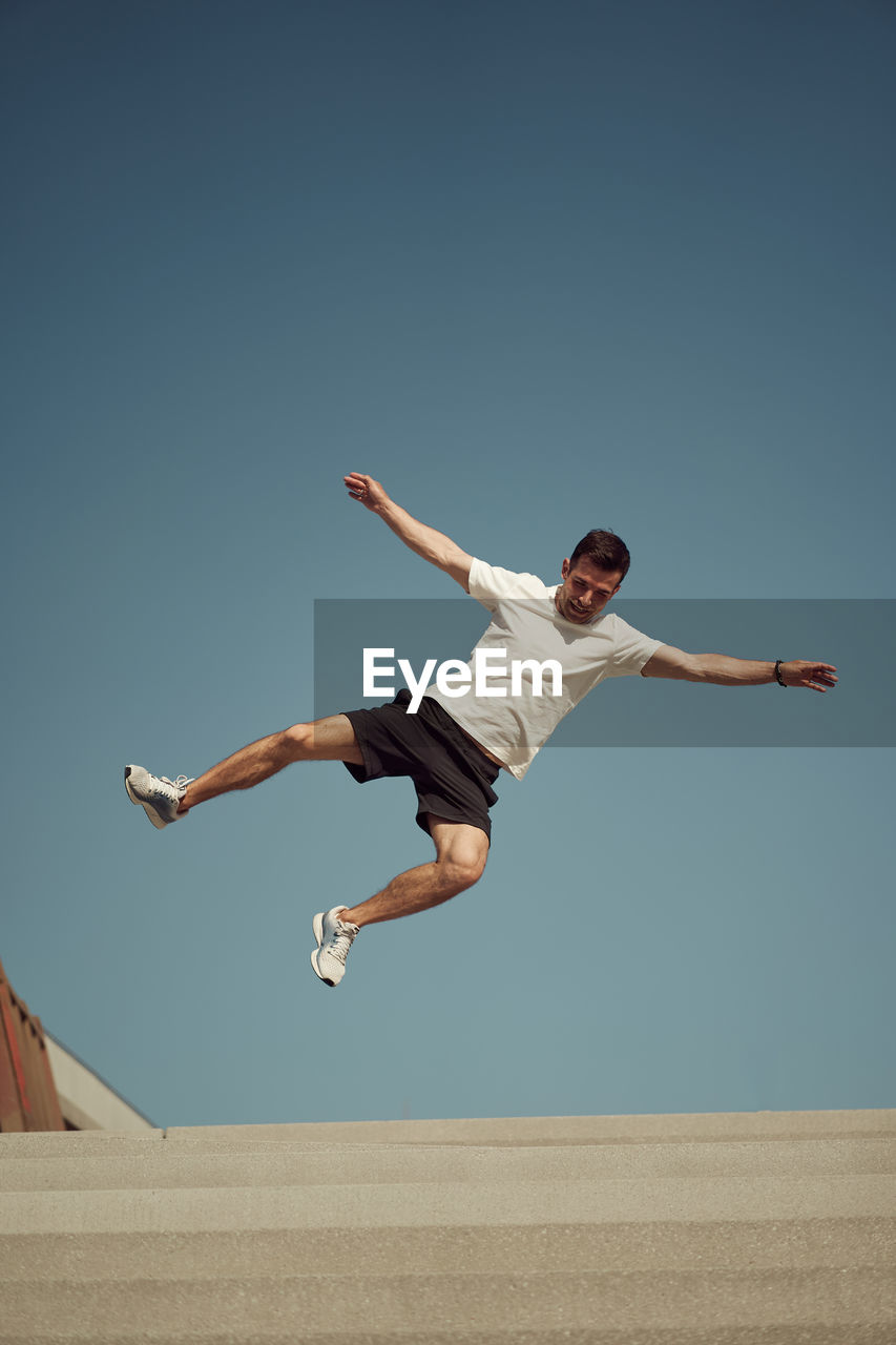 From below of active male jumping high and showing parkour stunt against blue sky in summer