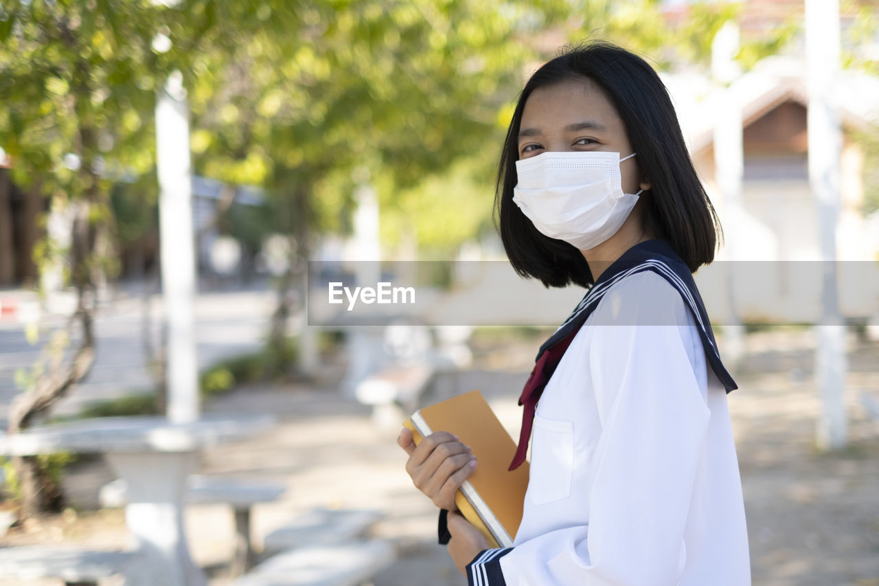 Portrait of girl wearing mask with book standing outdoors