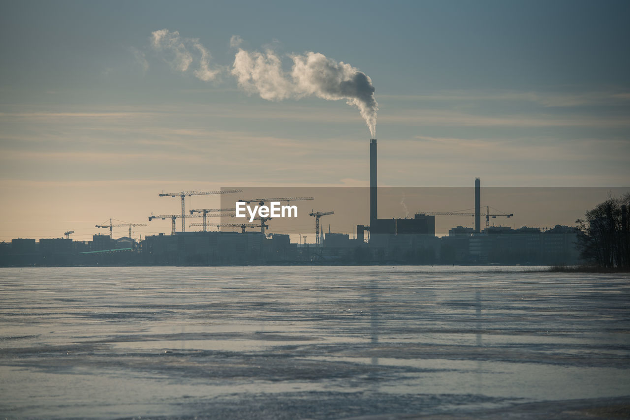 Lake by factory emitting smoke against sky during winter