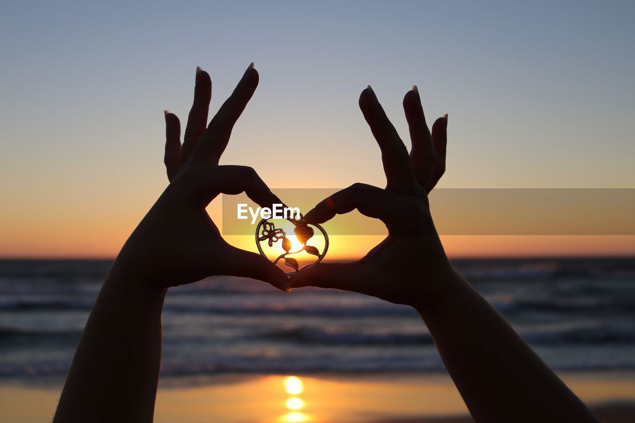 Cropped image of woman holding heart shape locket against clear sky during sunset