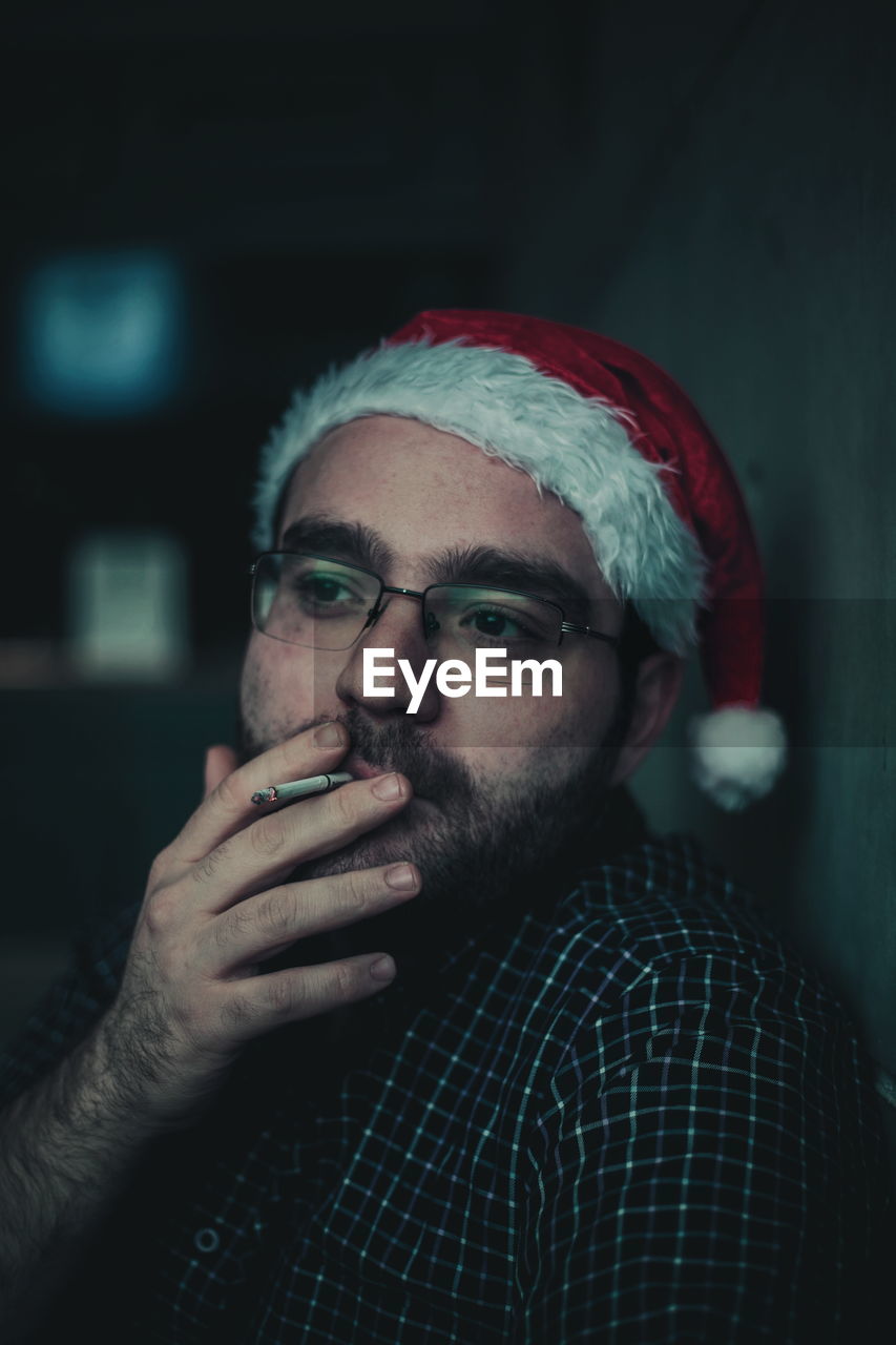 Thoughtful young man smoking cigarette while wearing santa hat at home