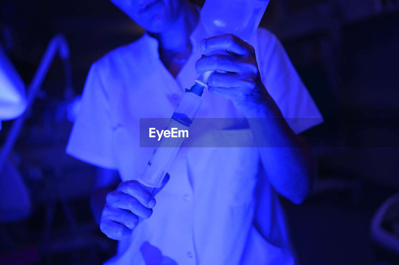 Cropped unrecognizable woman in medical uniform using syringe to take drug from bottle while standing near incubator in neonatal ward with blue illumination during night shift