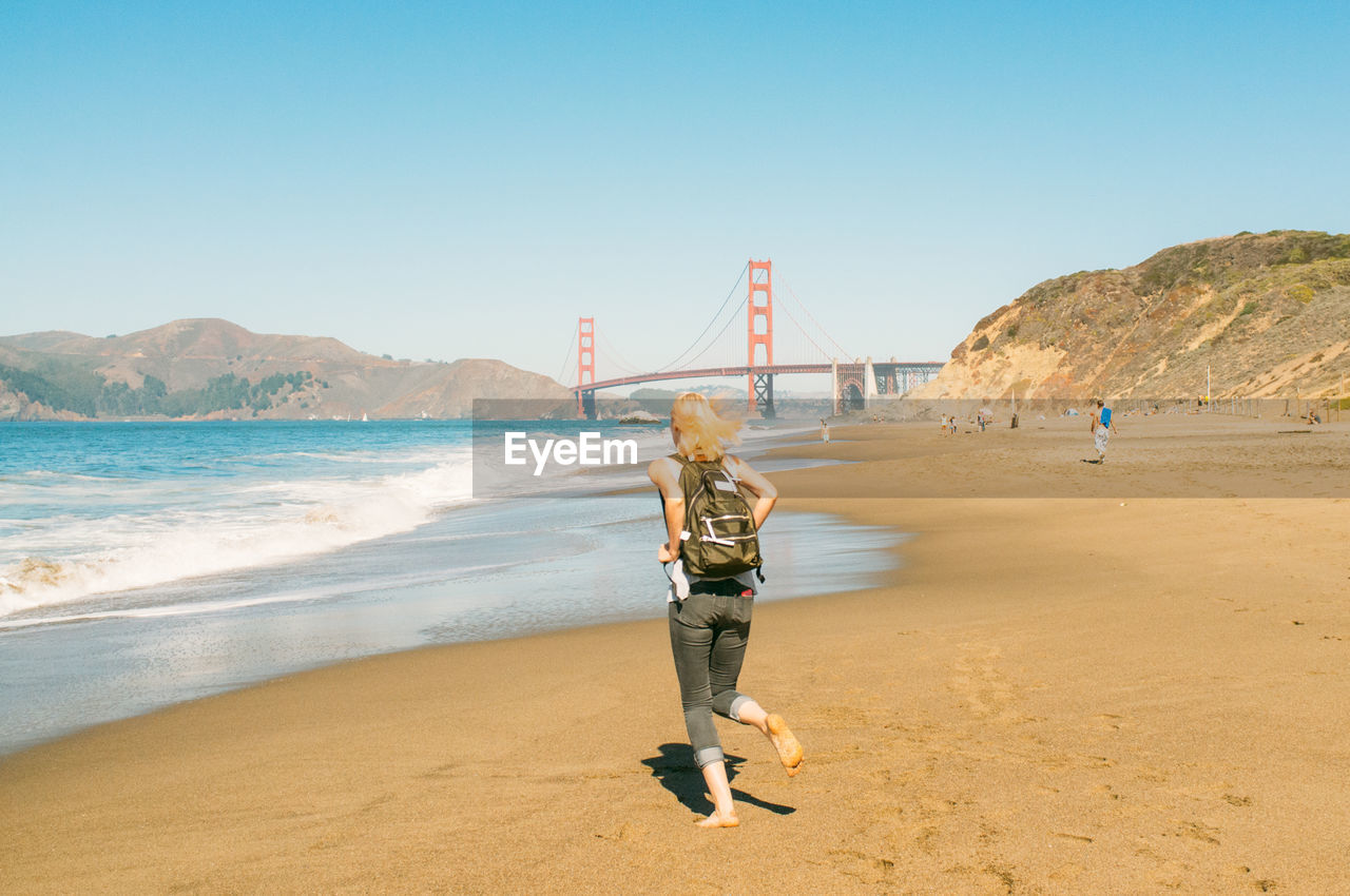 Full length rear view of woman running on beach by golden gate bridge against clear blue sky