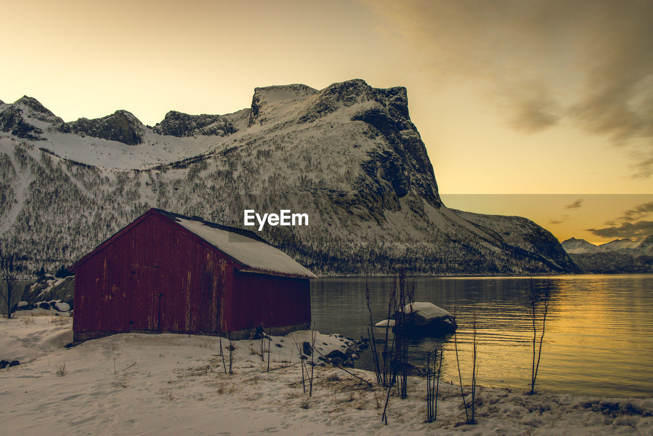 Wooden house on snow against sky during sunset