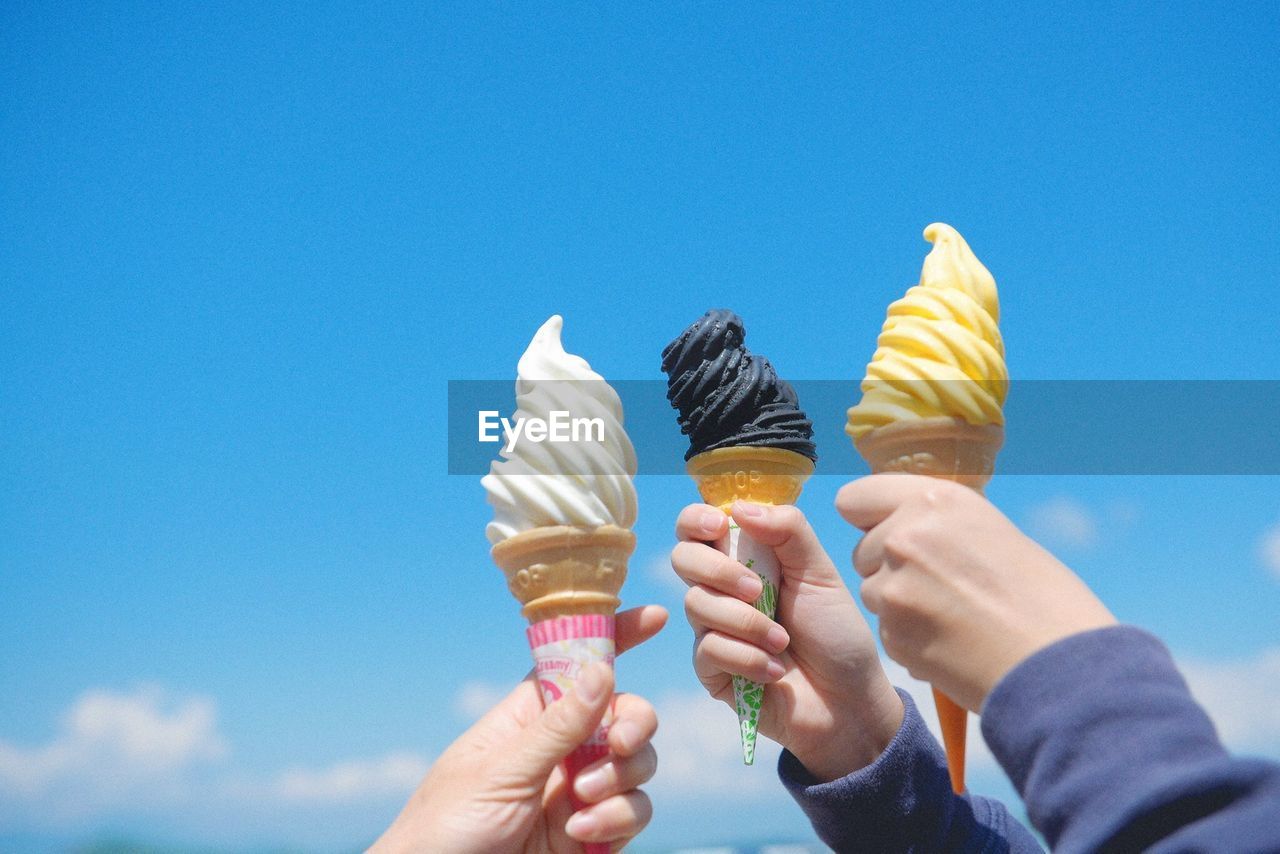 Cropped hands of people holding ice cream cone against blue sky