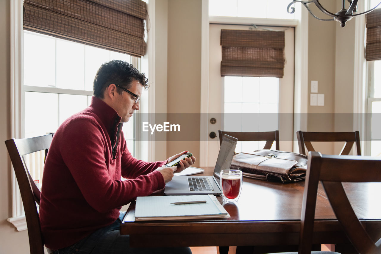 Man in glasses working from home using a computer at a dining table.
