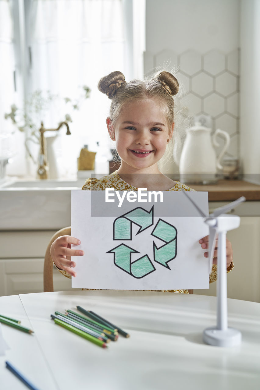 Smiling girl holding recycling symbol drawing on paper at table