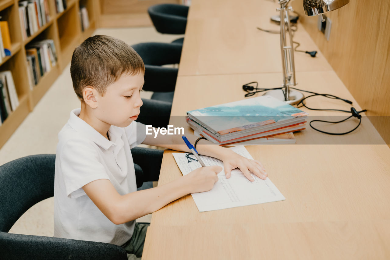 The boy sits in the library and writes in a notebook at the table. preparing for homework. 