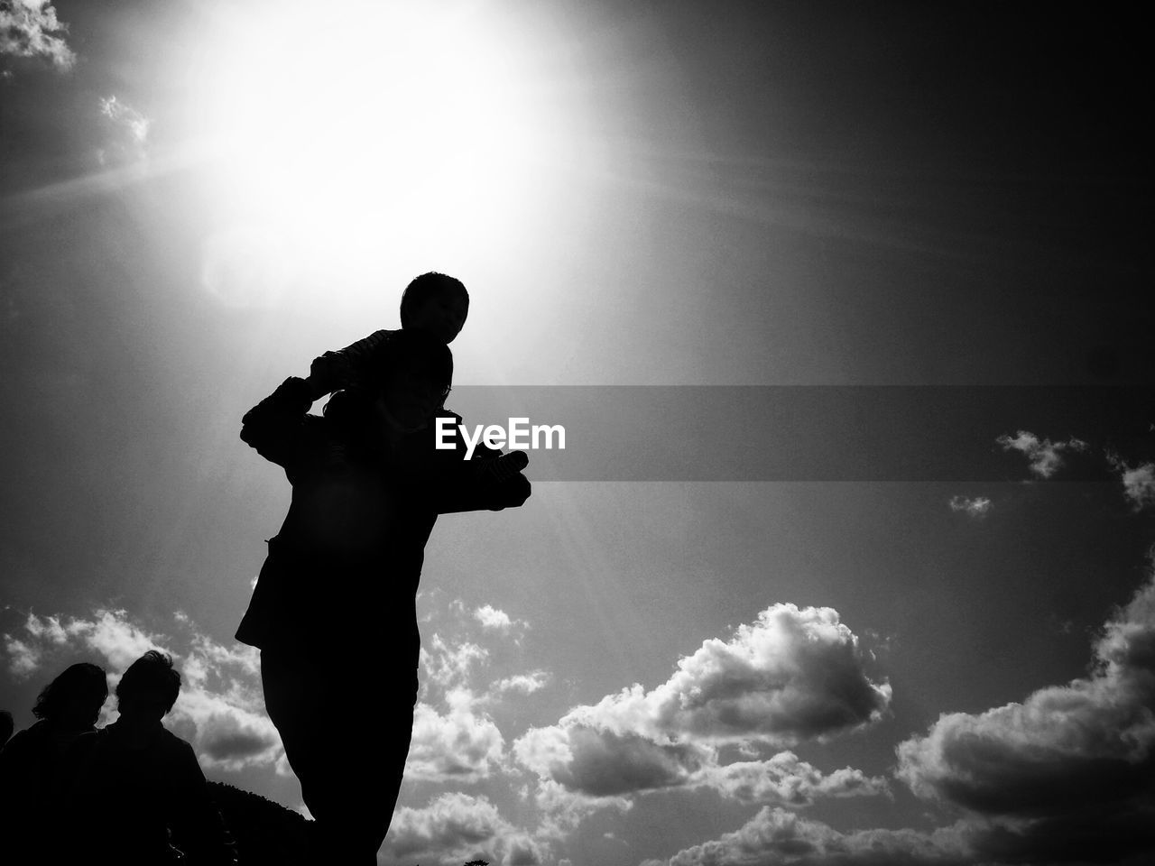 Low angle view of silhouette parent carrying child on shoulders against sky during sunny day