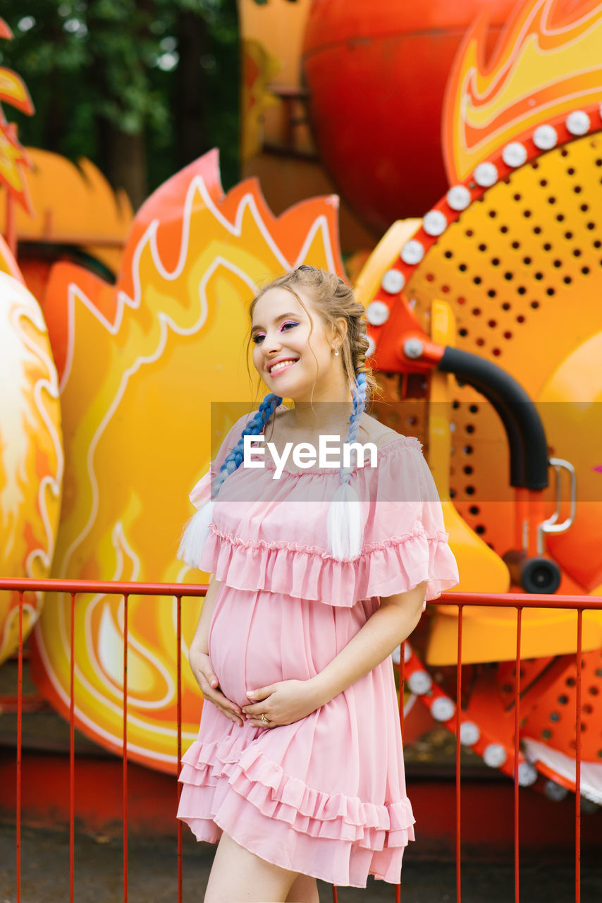 Cheerful pregnant young woman with blue pigtails and bright makeup walks in an amusement park