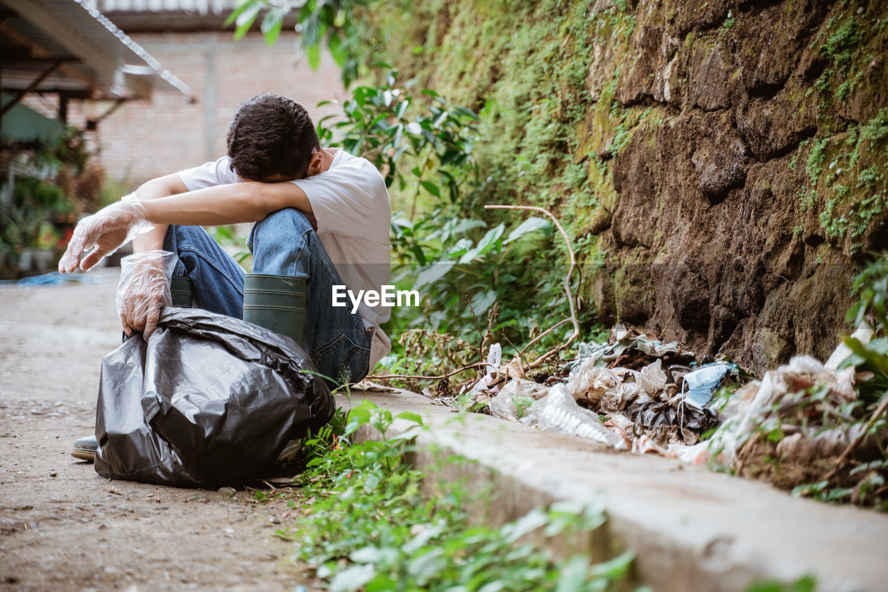 Tired man with garbage bag sitting roadside