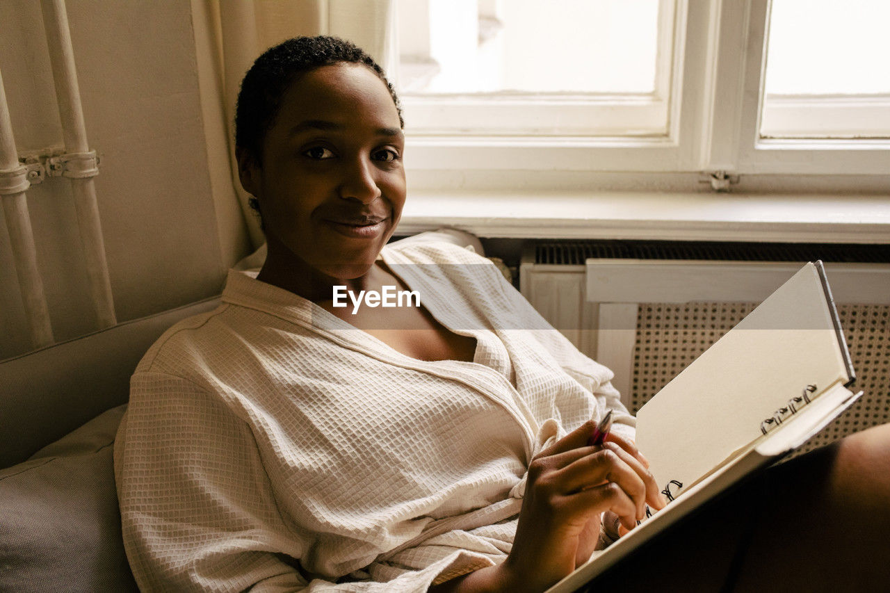 Portrait of smiling young woman with book in bedroom at home