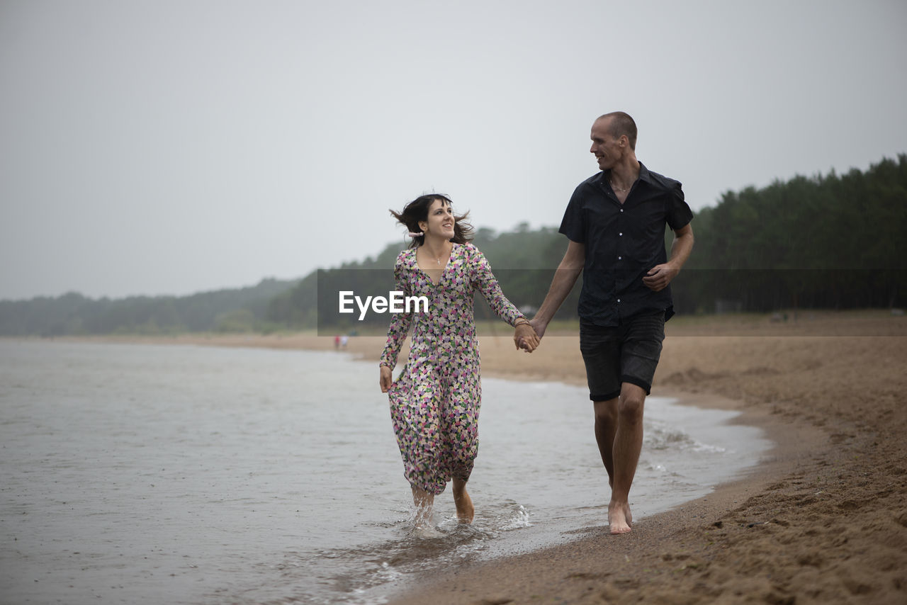 Young happy couple holding hands running along the sea shore ar rainy summer day