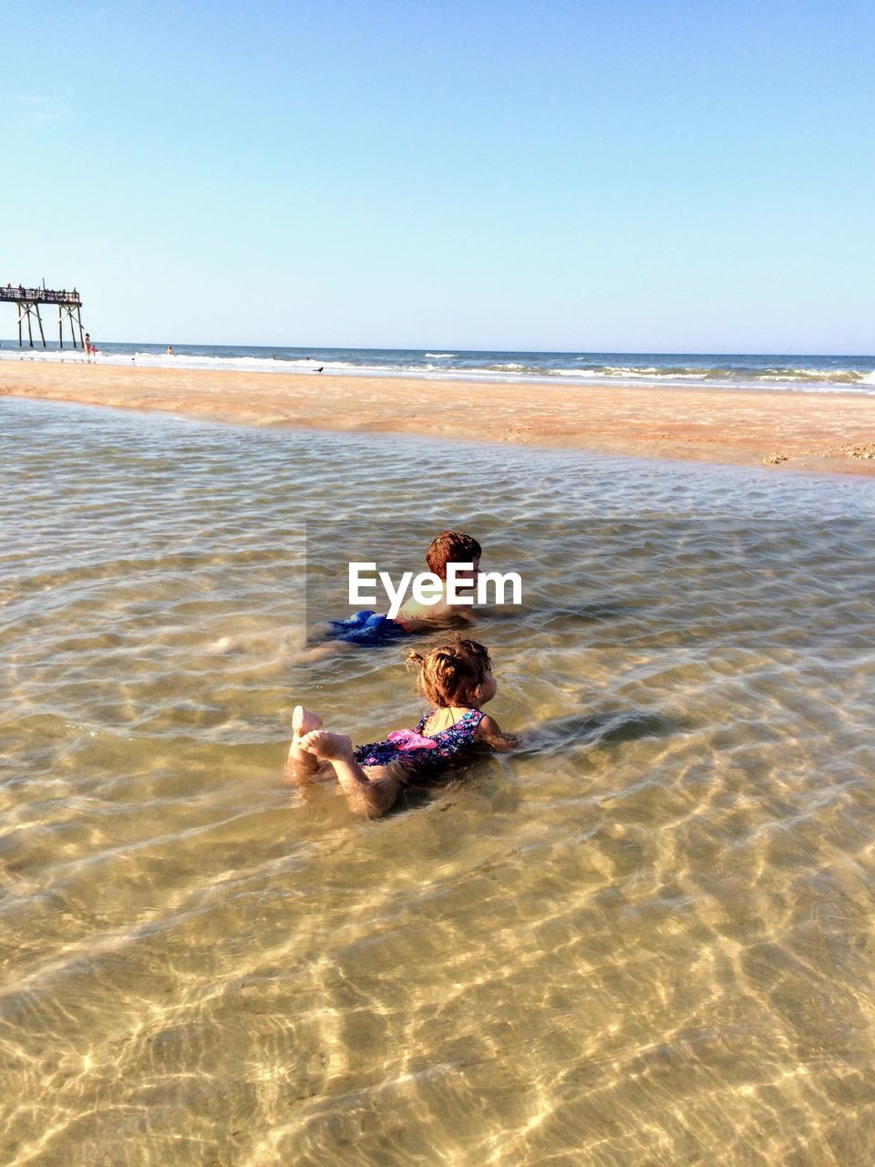 Siblings lying in tidal pool at beach against clear sky