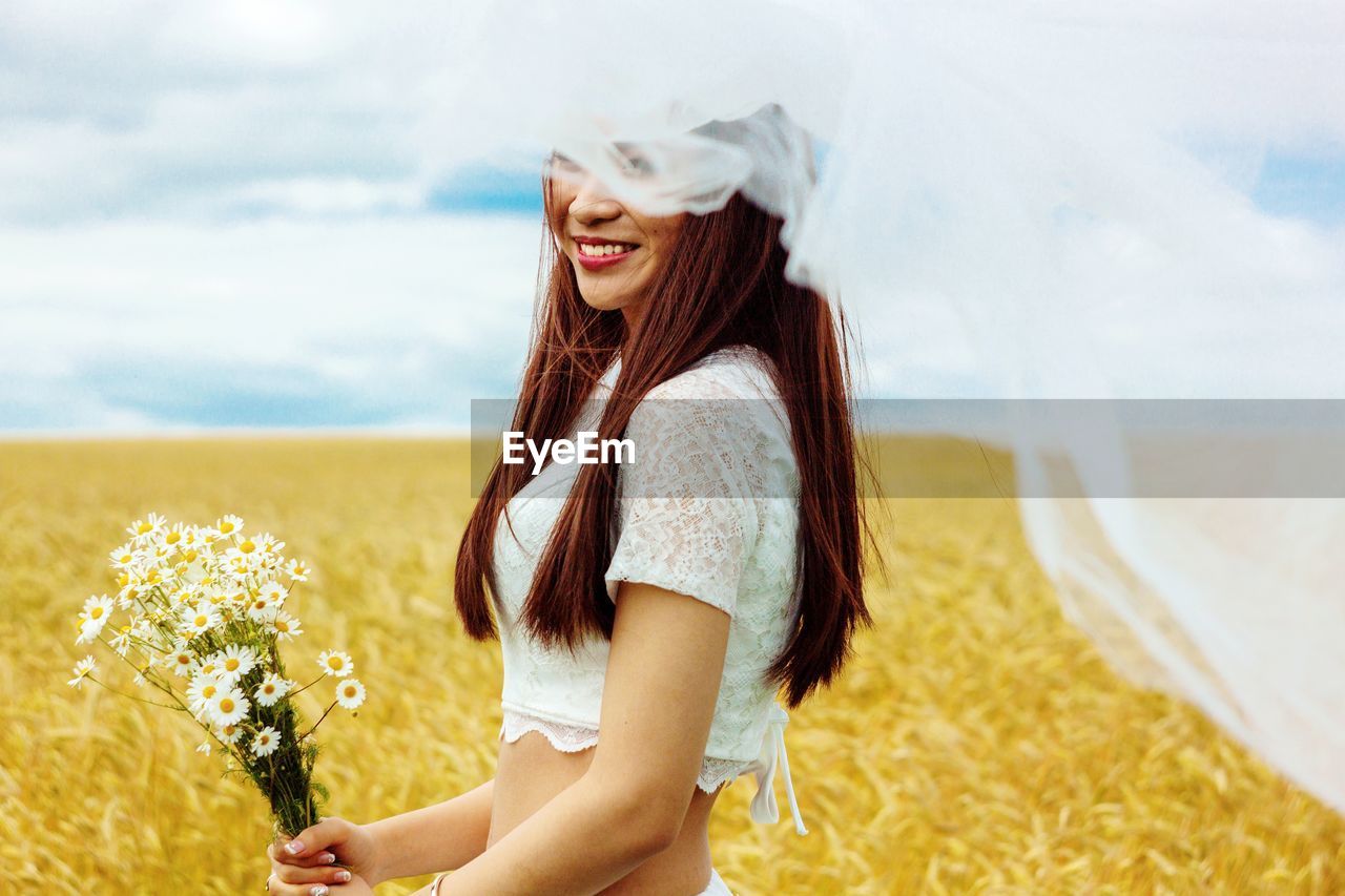Smiling bride in veil with flowers standing on agricultural field