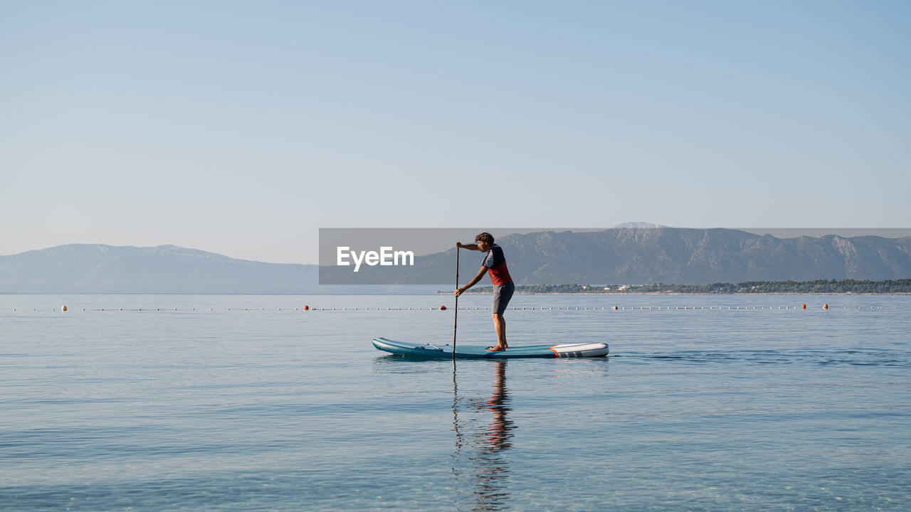 Man standing on paddleboard in sea against clear sky