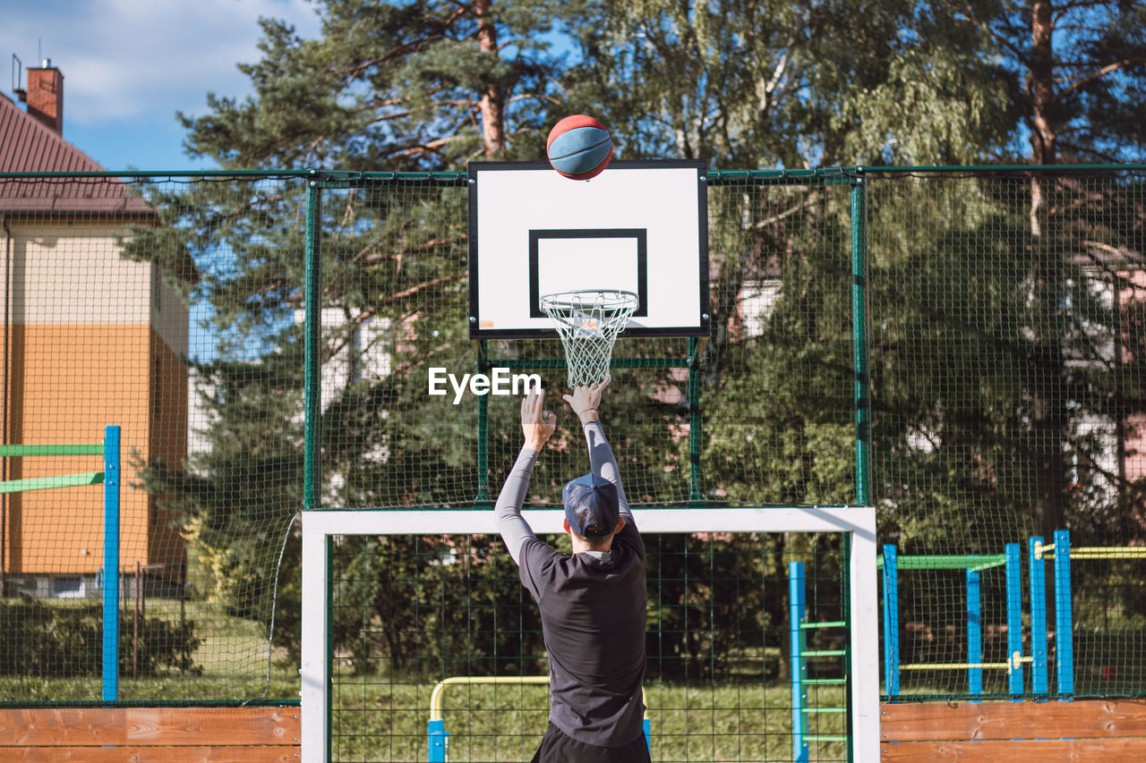 basketball, sports, basketball hoop, ball, player, playground, athlete, basketball - ball, basketball player, leisure activity, men, tree, competition, adult, day, lifestyles, nature, one person, full length, motion, throwing, net - sports equipment, young adult, vitality, city, plant, exercising, mid-air, arm, jumping, standing, team sport, outdoors, clothing, casual clothing, competitive sport, sports equipment, arms raised, person