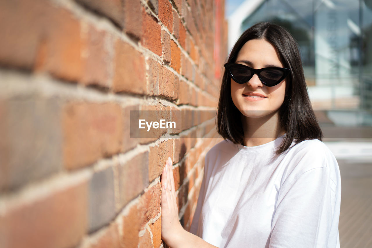 portrait of young woman wearing sunglasses standing against brick wall