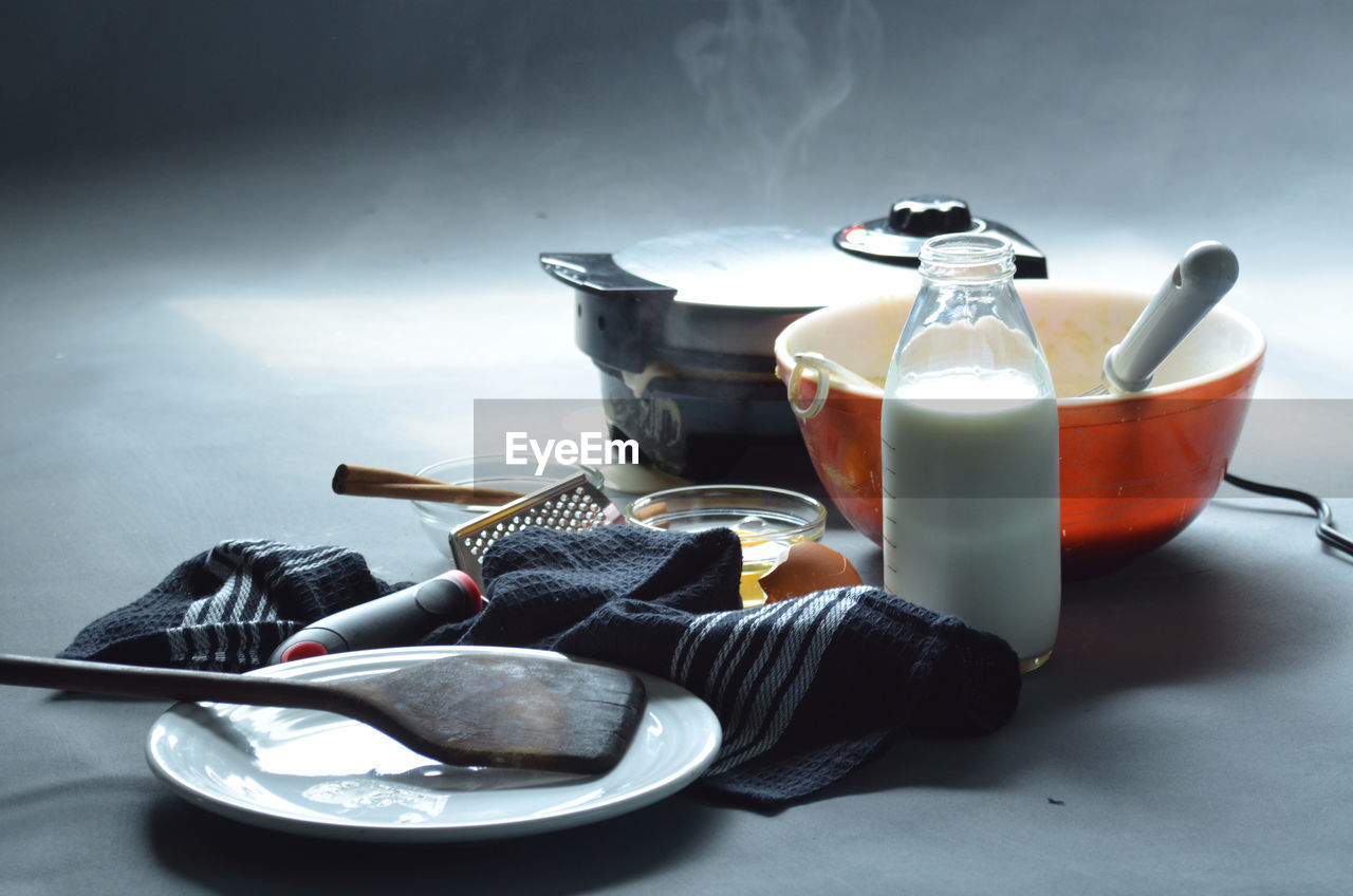 Steaming waffle iron with bowl of batter, bottle of milk, cinnamon sticks and cooking utensils