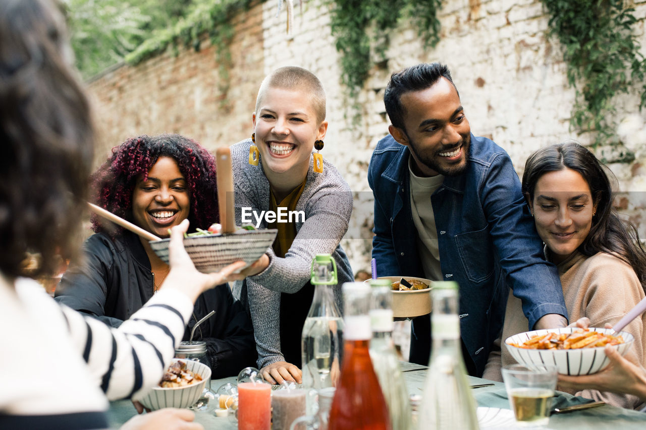 Young woman with toothy smile passing bowl to friend at dinner party