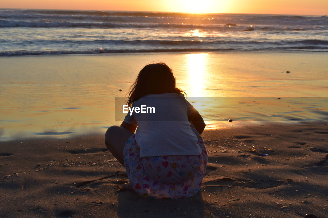 rear view of woman sitting at beach against sky during sunset