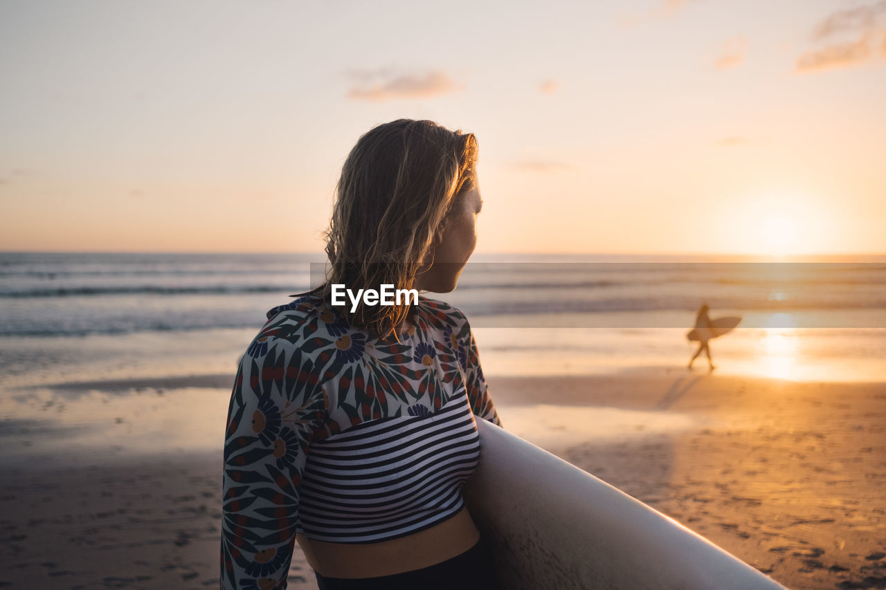 Mid adult woman with surfboard looking at sea during sunset at beach