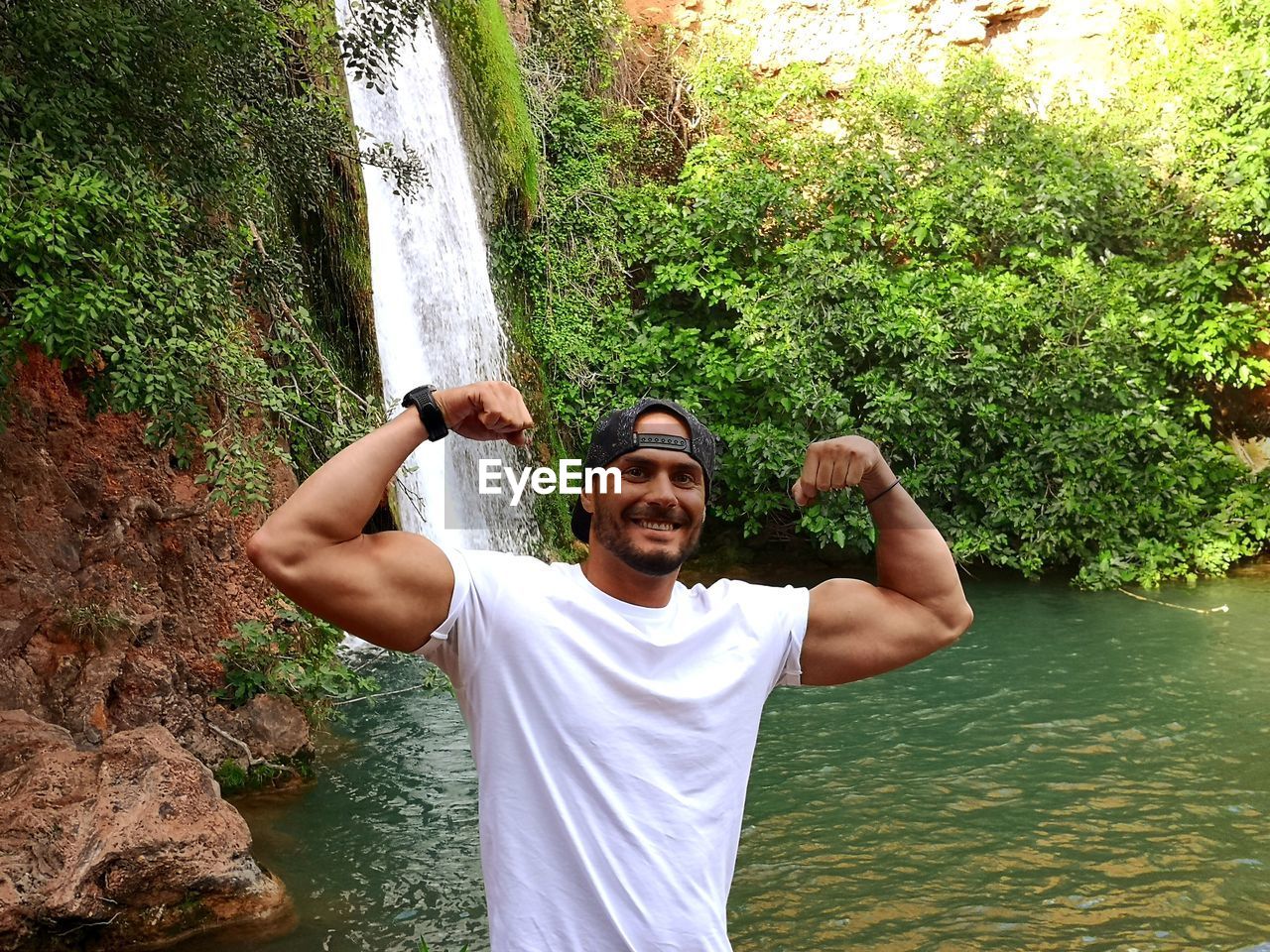 Portrait of smiling young man flexing muscles while standing against waterfall