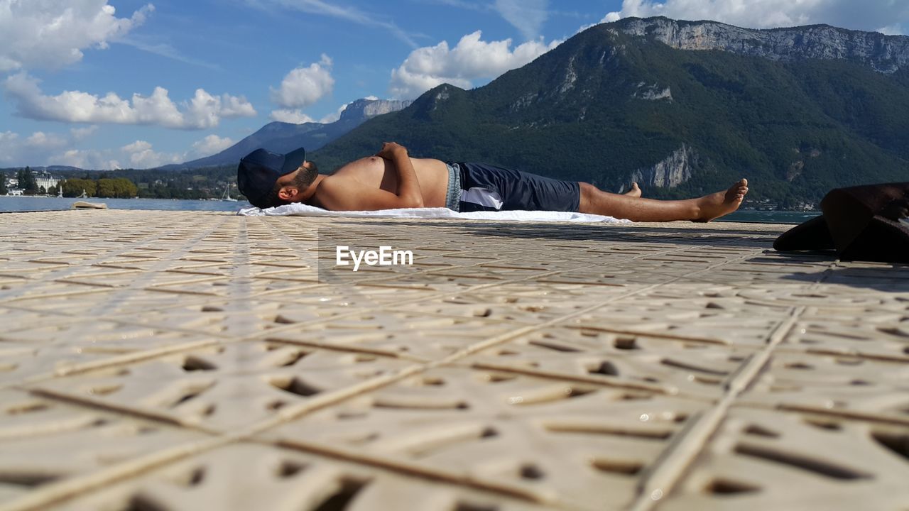 Surface level shot of shirtless man sleeping by lake against mountain