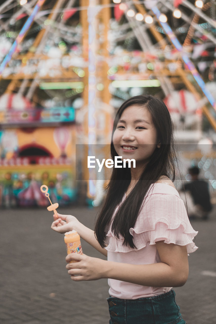 Portrait of smiling young woman holding bubble wand at amusement park