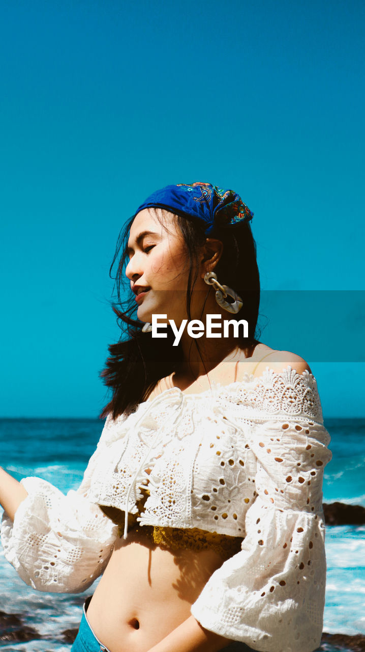 Beautiful young woman standing by sea against blue sky