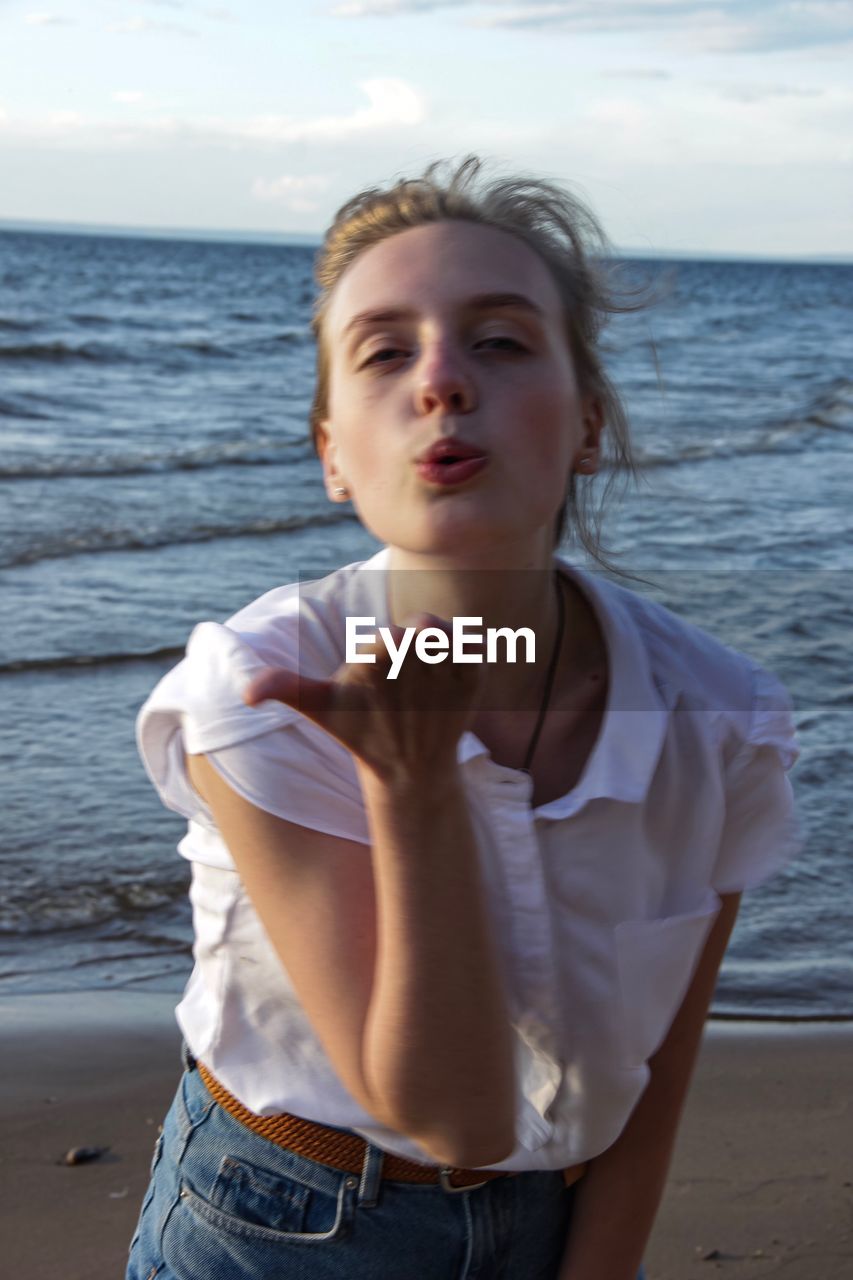 Portrait of young woman blowing kiss while standing at beach