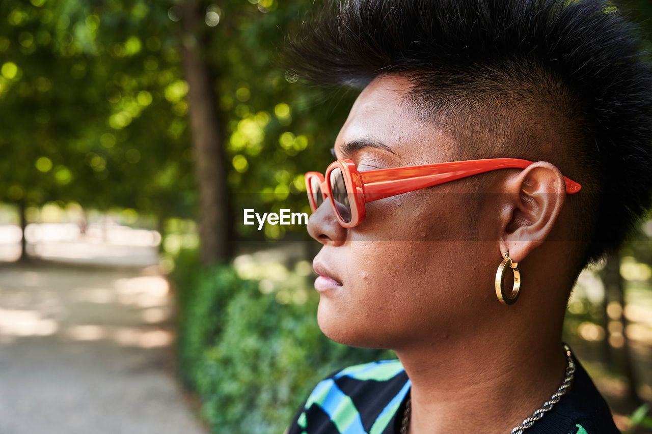 Filipino non binary person in stylish sunglasses with acne on face looking away while standing near tropical lush bush on summer day in park