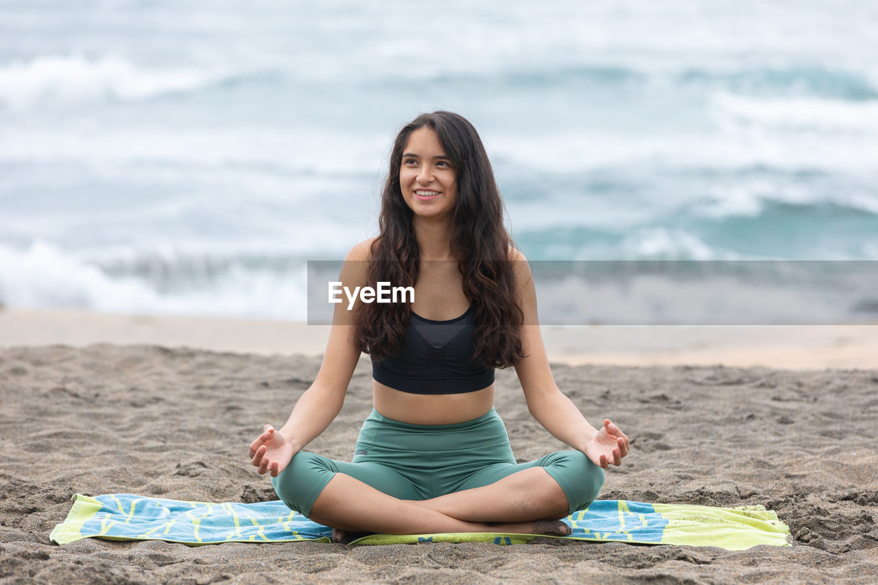 Content young ethnic woman sitting on seashore in padmasana pose during yoga practice