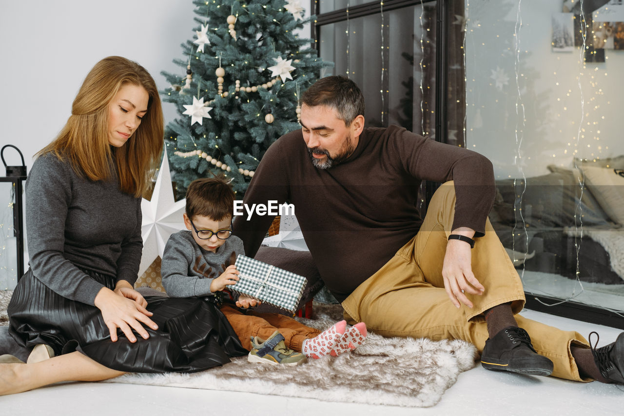 Family with child playing near christmas tree. child unpacking gifts, parents enjoy christmas tree