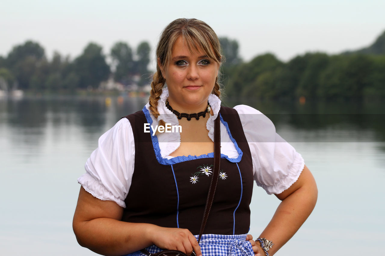 Portrait of smiling oktoberfest waitress standing with hand on hip against lake