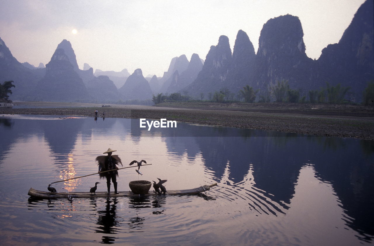 Man sailing boat with birds in river against mountains