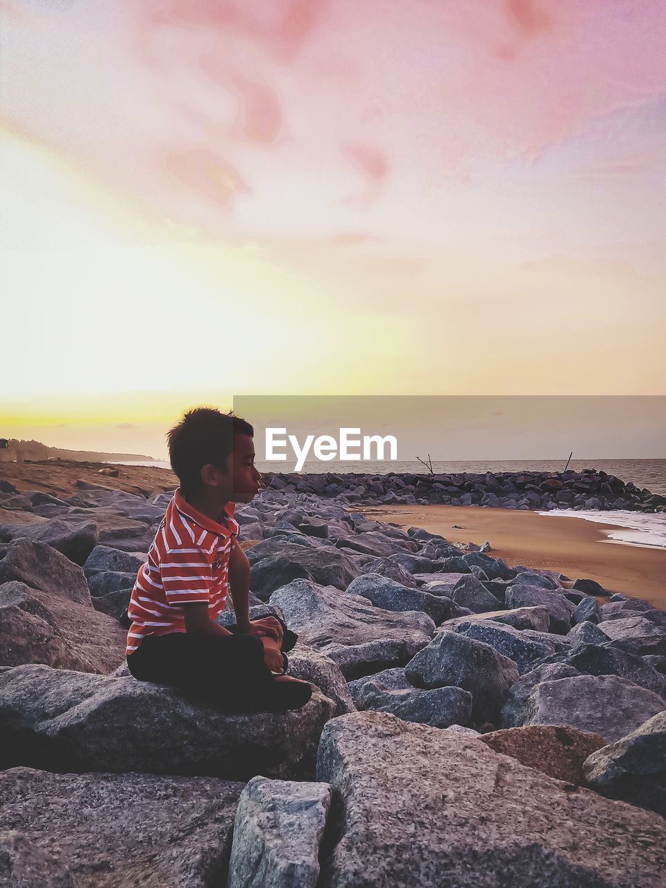 Side view of child sitting on rock at beach against sky during sunset