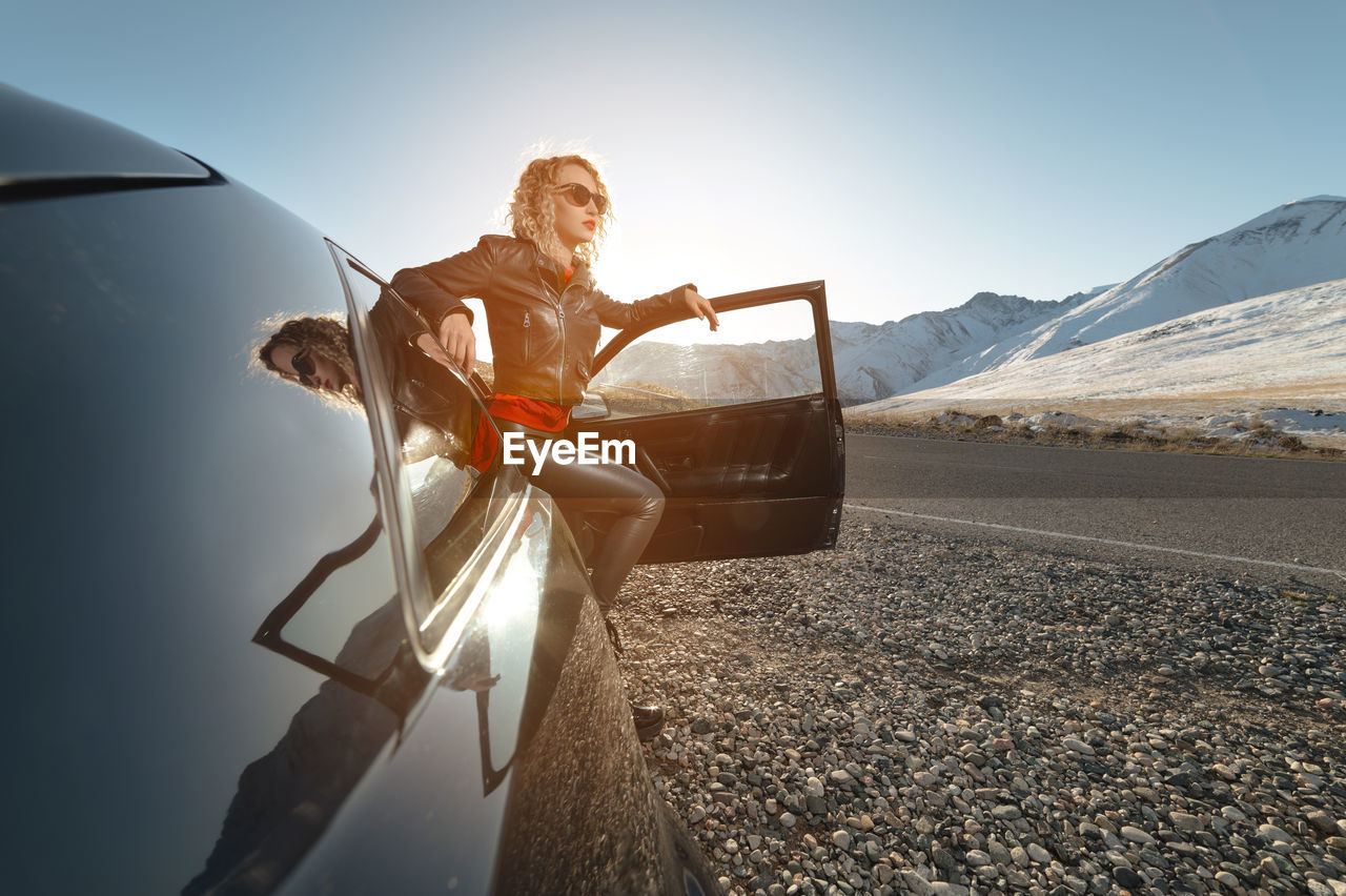 Fashion girl standing next to a retro sports car in the sun. stylish woman in a leather suit 