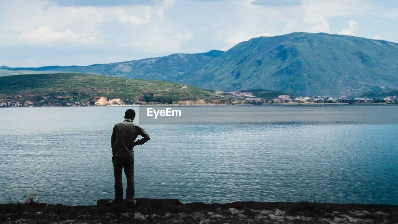 Rear view full length of man standing at lakeshore against mountain