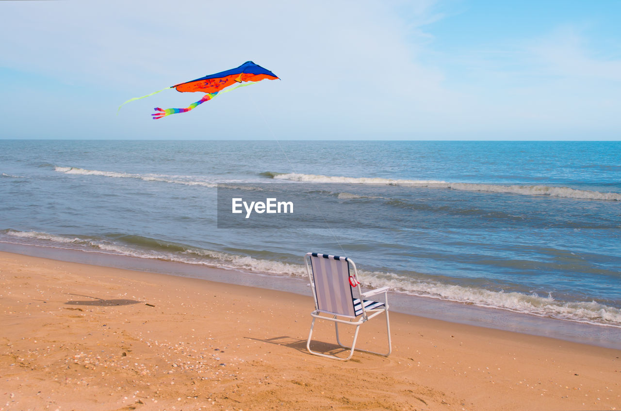 Kite flying over empty folding chair on shore at beach against sky