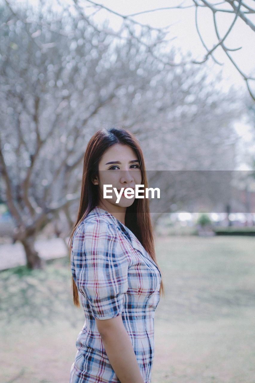 Portrait of young woman standing by bare trees in park