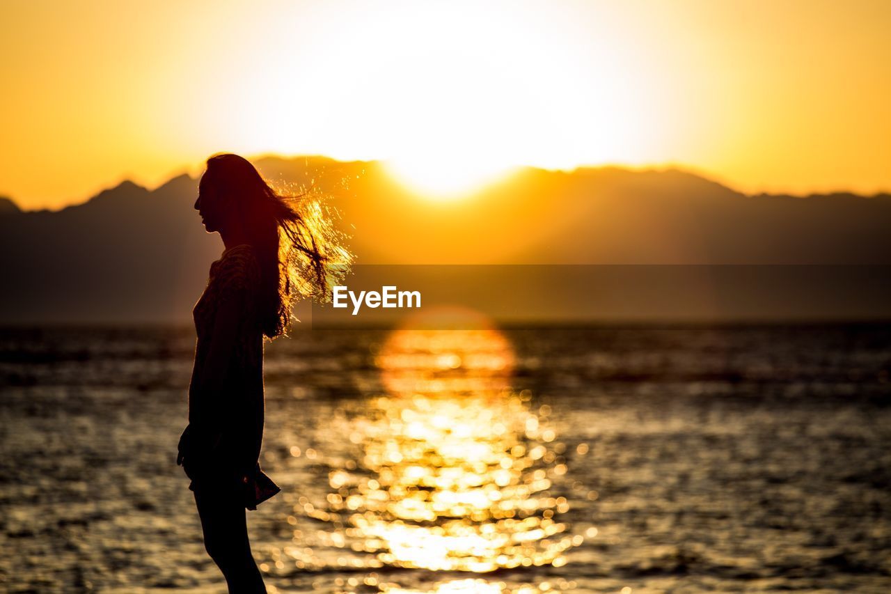 SIDE VIEW OF SILHOUETTE WOMAN STANDING BY SEA AGAINST ORANGE SKY