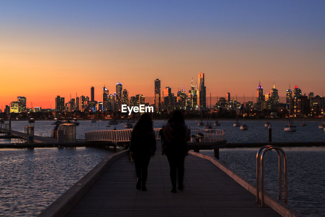 Friends walking on pier over sea against buildings during sunset