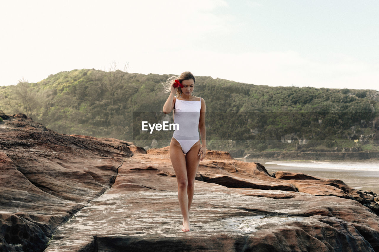 Seductive young woman wearing white swimsuit while standing on rock at beach