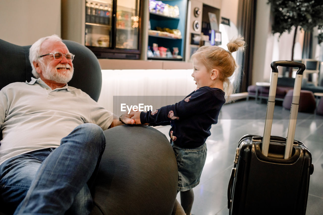 Side view of girl pulling grandfather sitting on armchair in hotel lobby