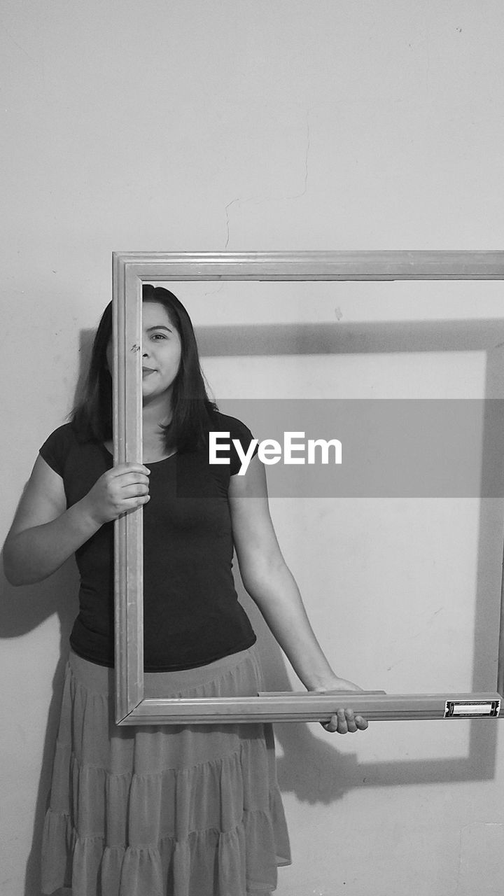 Portrait of woman holding frame while standing against wall