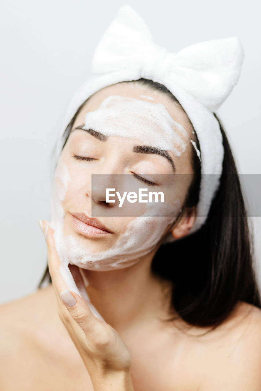 human face, body care, adult, skin, human head, women, portrait, one person, headshot, beauty treatment, young adult, indoors, skin care, eyes closed, beauty spa, studio shot, spa treatment, relaxation, wellbeing, facial mask - beauty product, hygiene, nose, human skin, close-up, beauty product, health spa, exfoliation, female, brown hair, clothing, front view, white background, cleaning, moisturizer, bathroom, lifestyles, veil, headband, domestic bathroom, towel, make-up, applying
